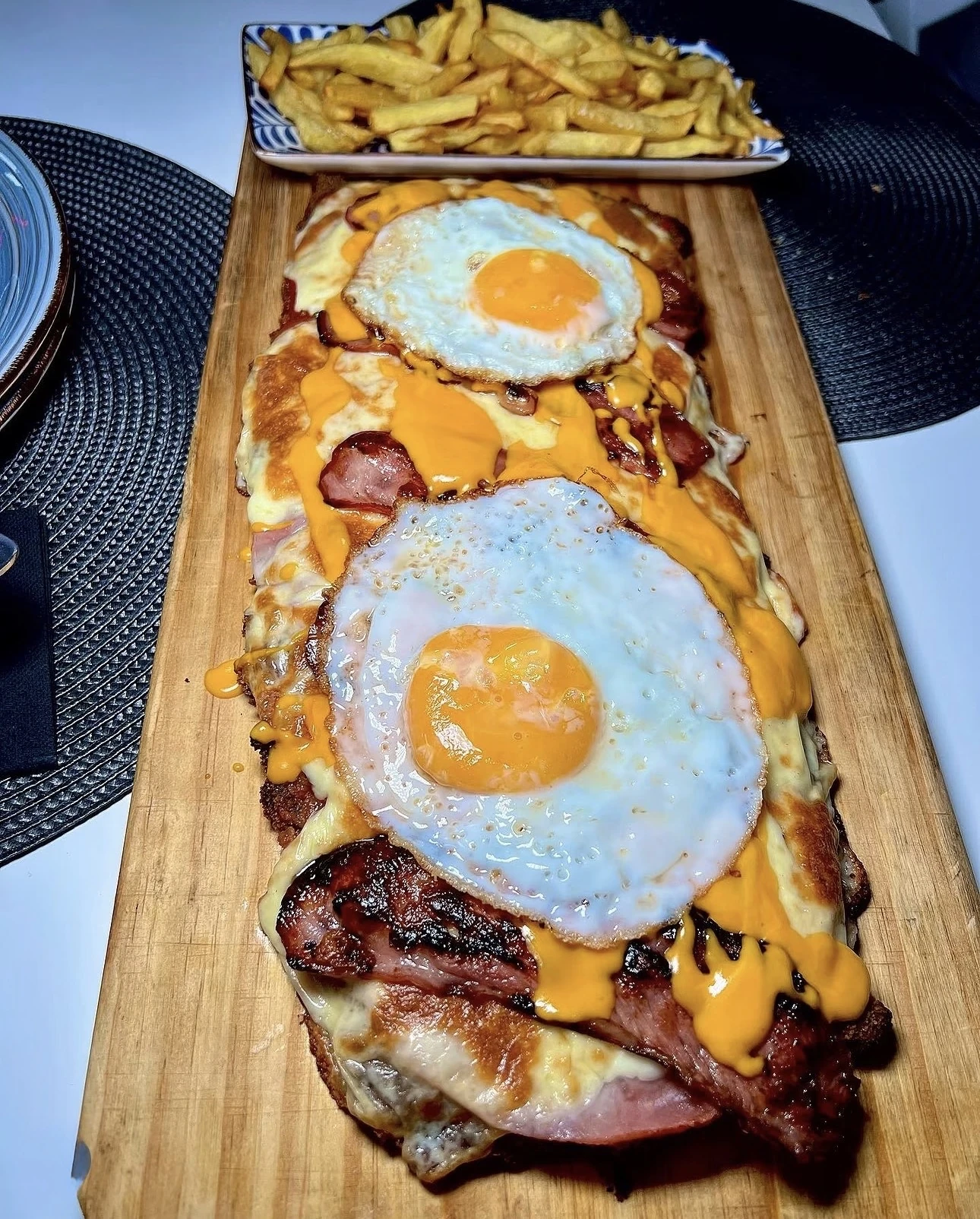 Milaneson de entraña sobre tabla de madera con dos huevos fritos, cheddar y beicon, con fuente de patatas fritas