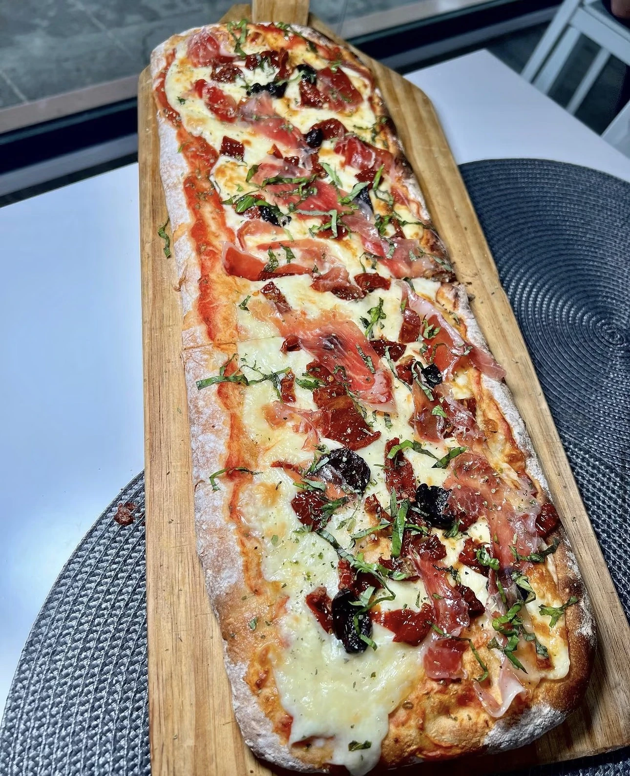Pizza rectangular de jamón, tomate seco, olivas negras y albahaca sobre pala de madera